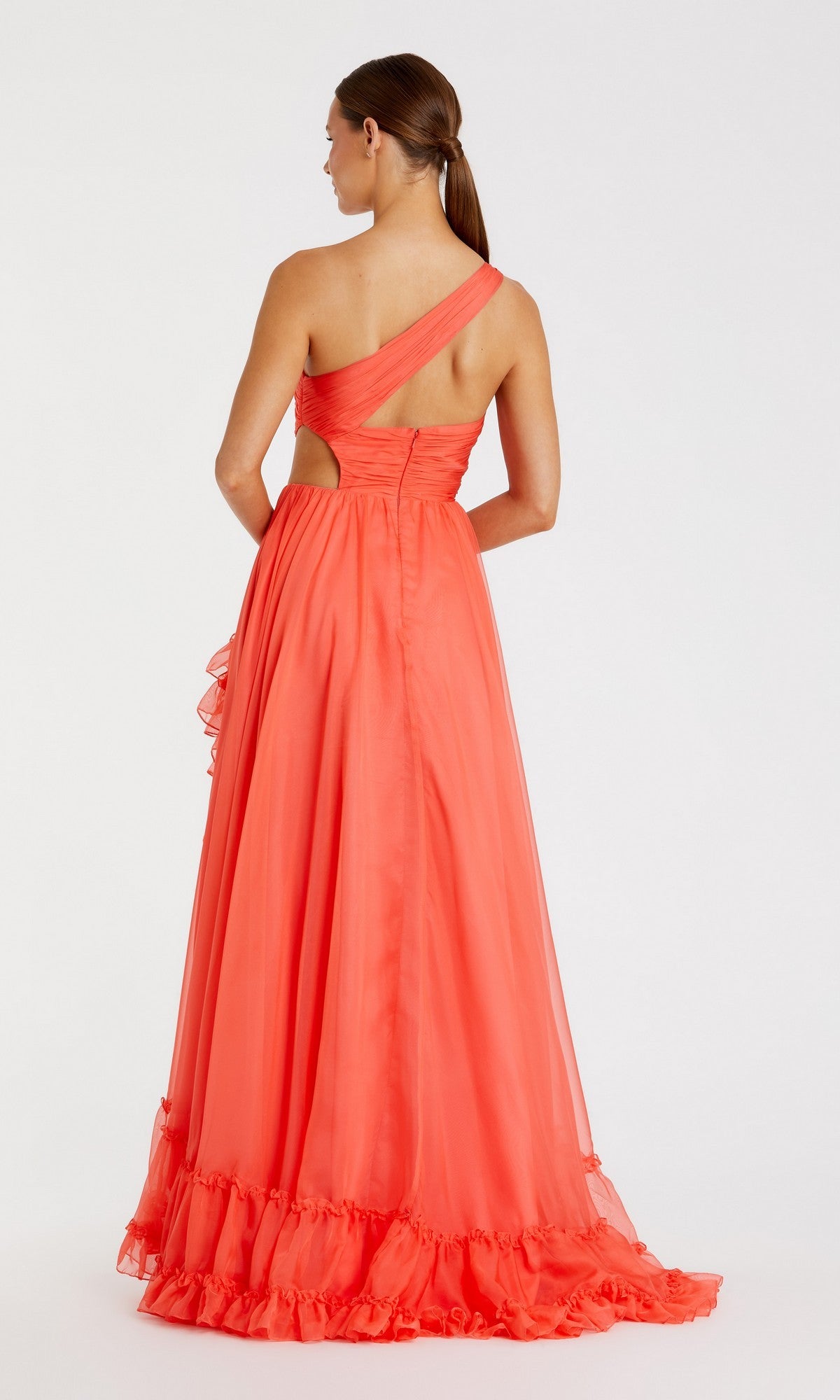 A woman stands facing away, showcasing the Mac Duggal 68476 long formal dress in coral, featuring a one-shoulder design with a ruffled hem and cutout detail on the back, set against a plain white background.